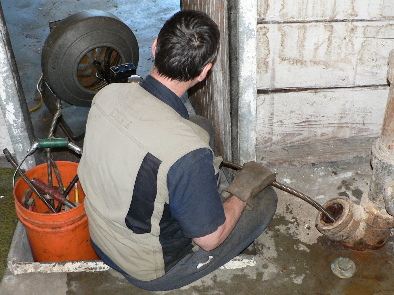 A worker crouched down using a drain cleaning machine to clear a clogged pipe in an industrial or workshop setting.