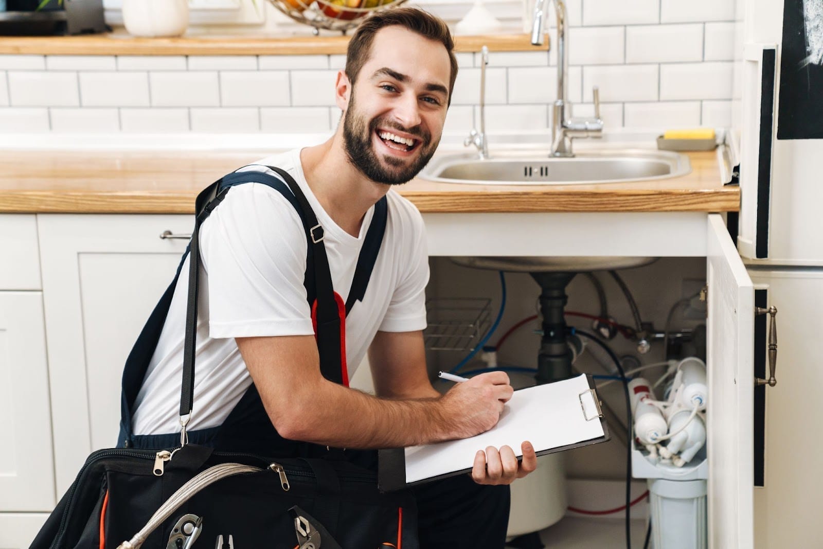A cheerful handyman with a clipboard preparing to work under a kitchen sink.