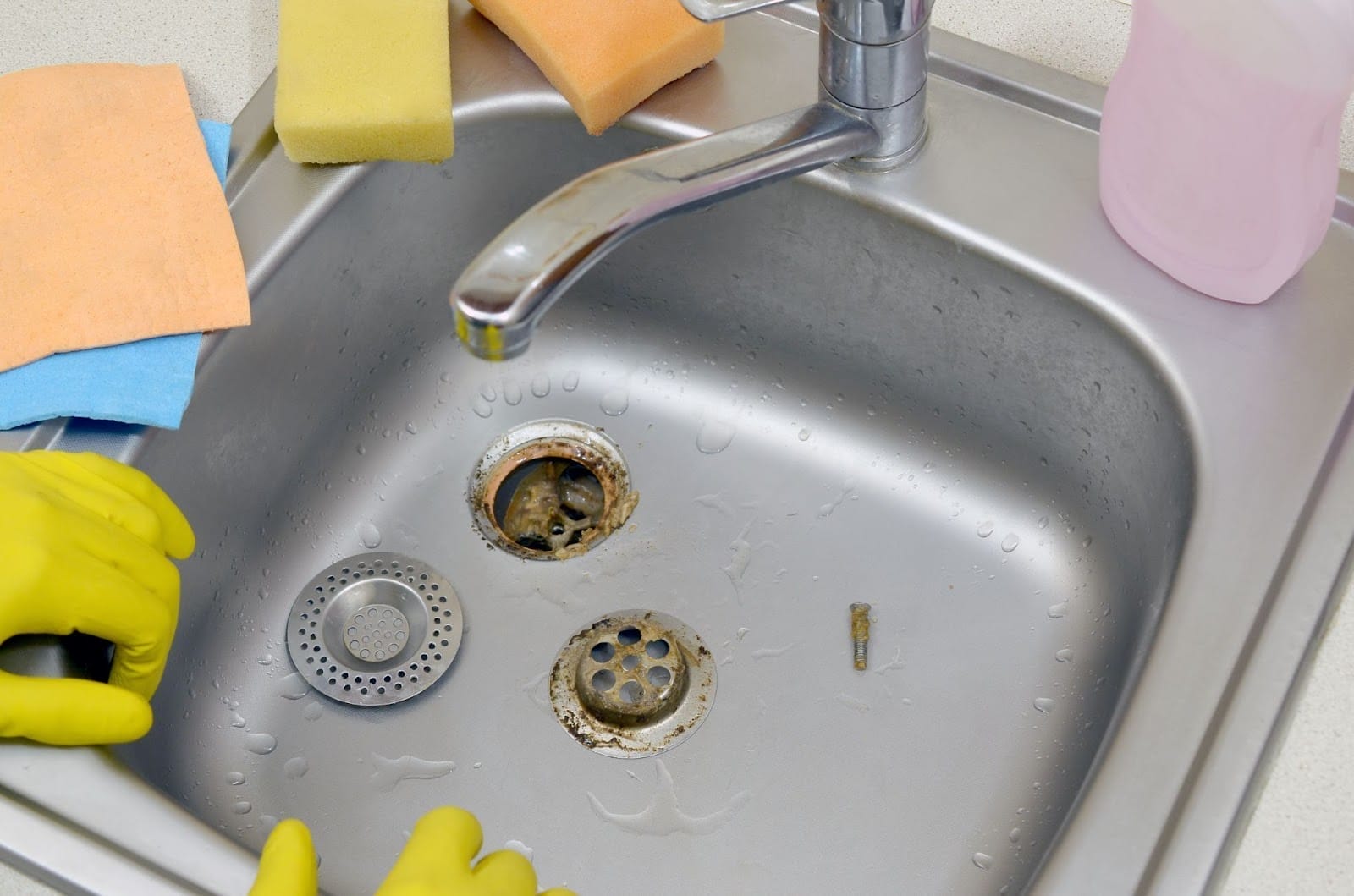 A person wearing yellow rubber gloves is about to clean a dirty stainless steel kitchen sink with various cleaning supplies nearby.