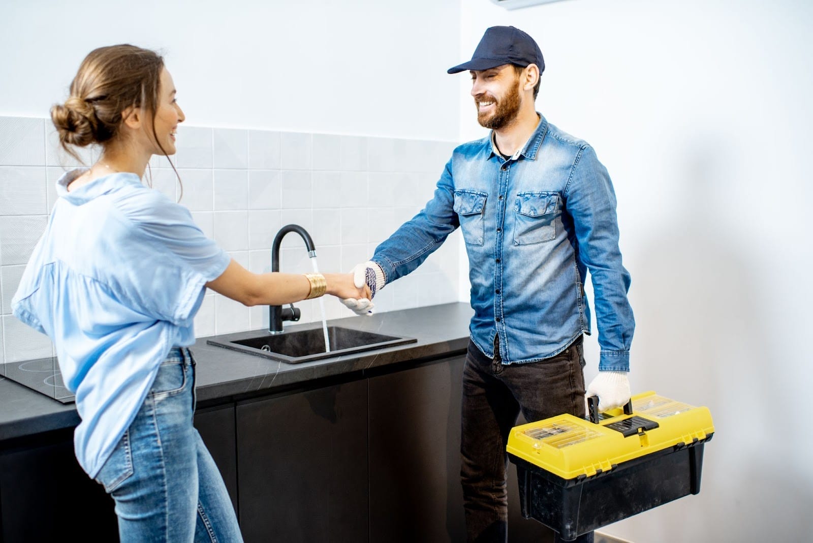 A smiling handyman in a denim jacket hands a glass of water to a cheerful woman in a kitchen, with a toolbox placed on the countertop, suggesting a friendly interaction possibly after a successful home repair.