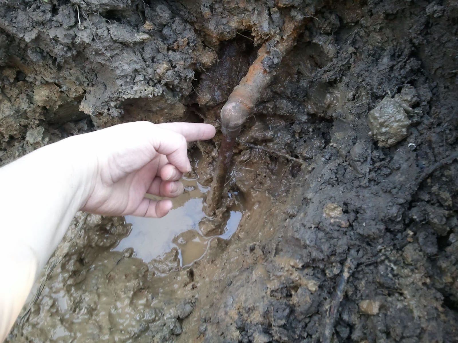 A hand pointing at a rusty pipe exposed within a muddy excavation site.