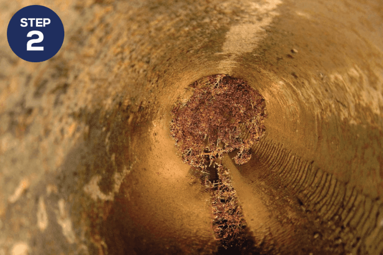 A close-up view inside a pipe with a significant accumulation of debris and grime. The image highlights the need for cleaning or maintenance. On the top left corner, a dark blue circular label with the text "STEP 2" is visible.
