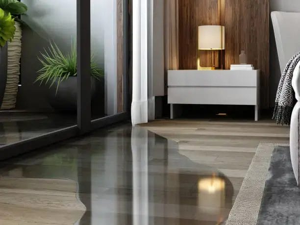 A puddle of water on a glossy wooden floor in a modern bedroom, reflecting a lit lamp.