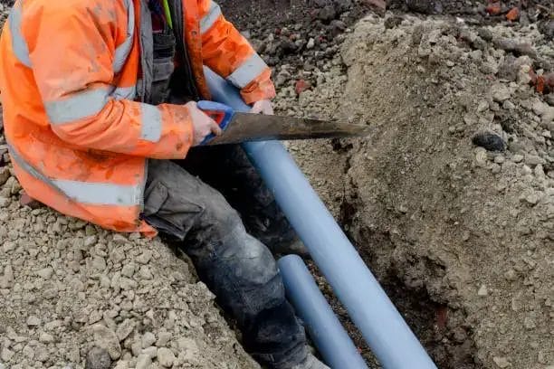 Worker in orange safety vest cutting blue pipes in a trench.