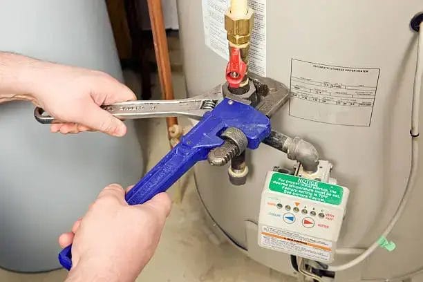 Hands using two pipe wrenches to tighten or loosen a connection on a residential water heater, focusing on the inlet valve and surrounding plumbing.