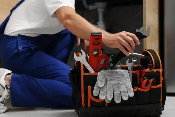 A worker in blue overalls kneels with a tool bag containing wrenches, a level, and gloves near a sink pipe.