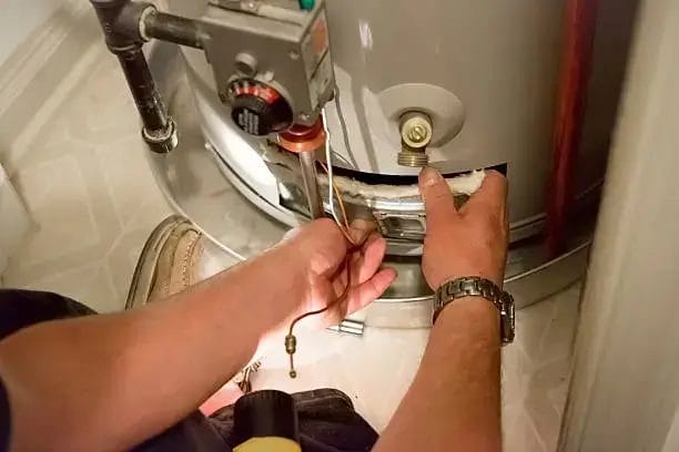 A technician uses a screwdriver to perform maintenance on a water heater, accessing components behind a small panel near the bottom of the unit.